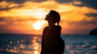 Silhouette of young woman at sunset, head tilted back against dramatic orange sky and ocean backdrop. Peaceful meditation pose captures serenity and freedom by seaside.