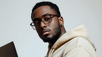 Young African American male professional wearing glasses and cream hoodie looking at laptop screen with focused expression against light background.