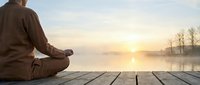 Man meditating in lotus position on wooden dock at sunrise over misty lake. Peaceful morning yoga practice for mindfulness and spiritual wellness.