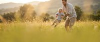 Father and toddler son playing together in golden sunset meadow, family bonding time outdoors in nature with warm sunlight and mountain landscape background.