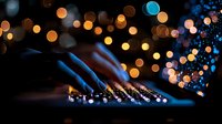 Illuminated computer keyboard with blurred bokeh lights creating magical atmosphere, close-up shot in dark environment with blue and orange glowing circles.