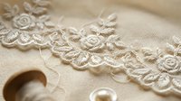 Delicate cream lace trim with floral embroidery and pearl beads on fabric, suitable for wedding dress design, bridal fashion, and textile decoration projects.