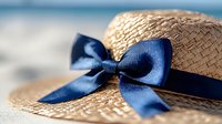 Woven straw sun hat with navy blue ribbon bow on sandy beach, summer vacation accessory for tropical getaway and seaside relaxation.