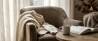 Cozy living room interior with comfortable armchair, knitted throw blanket, coffee mug and open book on wooden table creating warm hygge atmosphere for relaxation.