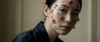 Young Asian woman with colorful floral face paint stickers on cheeks and forehead wearing dark green shirt in soft natural lighting for beauty concept.