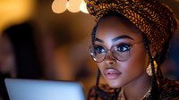 Young African woman in traditional headwrap and glasses working on laptop in evening, illuminated by warm ambient light. Professional remote work concept.
