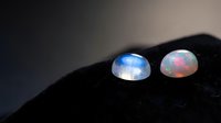 Two luminous rainbow moonstone cabochons displaying blue and pink adularescence on dark textured surface for jewelry making and gemstone collection.