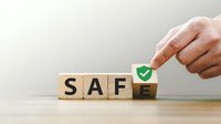 Hand placing green checkmark on wooden blocks spelling SAFE, representing security verification and safety assurance in business operations.