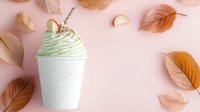 Mint green whipped cream dessert in textured white cup with cookie garnish on pink background surrounded by scattered autumn leaves and petals.