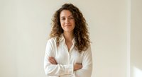 Professional young woman with curly hair in white shirt smiling confidently with crossed arms against neutral background for business content.