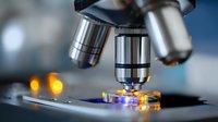 Close up view of microscope lenses examining illuminated specimen on glass slide in laboratory setting with blurred bokeh lights in background.