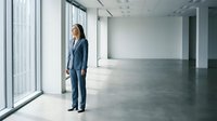 Young professional woman in business suit standing in empty modern office space with large windows and natural light, ready for corporate environments.