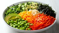 Fresh colorful poke bowl with edamame, avocado, shredded carrots, seaweed, quinoa and green herbs arranged in sections, topped with sesame seeds.
