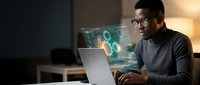 African American man working on laptop with holographic data visualization and digital interface projections in modern office environment at night.