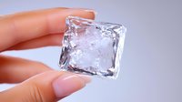 Clear ice cube held between fingers with french manicure against purple background, showing transparency, melting texture and cold refreshing effect.