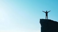 Silhouette of person standing with raised arms on mountain cliff edge against blue sky background, representing freedom, achievement and success.