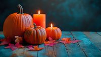 Orange pumpkins and glowing candles with red autumn maple leaves on rustic wooden table against dark teal background create cozy Halloween atmosphere.