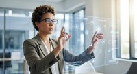 Professional mixed-race businesswoman with curly hair and glasses interacting with futuristic holographic interface in modern office environment.