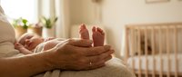 Tender moment of parent holding newborn baby feet in cupped hands with nursery crib in soft natural light background for family and parenting concepts.