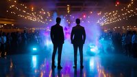 Two male silhouettes standing against colorful stage lights and crowd at concert venue with string lights overhead creating atmospheric nightclub party ambiance.
