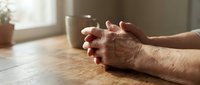 Elderly hands clasped in prayer on wooden table with coffee cup in soft morning light creating peaceful spiritual moment for meditation and reflection.