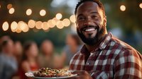Cheerful African American man holding plate of food at outdoor evening party with string lights, wearing plaid shirt, genuine smile at social gathering.