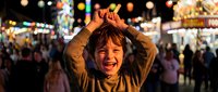Young Asian boy making playful gesture with hands on head at colorful night carnival with bright lights and crowd in background creating joyful atmosphere.