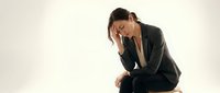 Stressed businesswoman in dark suit sitting with head in hands showing workplace burnout and mental health challenges on neutral background.