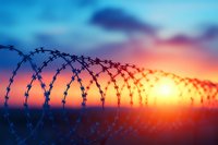 Silhouette of razor wire fence against dramatic sunset sky with orange and blue colors, symbolizing border security and restricted access. Suitable for security concepts.