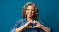 Smiling mature Caucasian woman with curly gray hair making heart shape with hands wearing denim shirt on blue background for wellness campaigns.