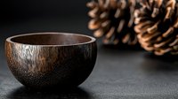 Handcrafted dark wooden bowl with natural textured surface against black background, pine cones creating rustic atmosphere. Minimalist eco-friendly tableware.