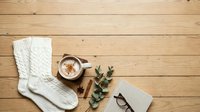 Cozy winter flatlay with white knit socks, warm beverage in ceramic mug, cinnamon sticks, eucalyptus sprigs, eyeglasses and open book on wooden surface.
