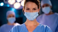Female medical professional in surgical mask and blue scrubs standing in operating room with blurred team members in background, dramatic lighting creates medical atmosphere.