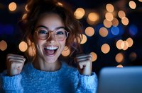 Young mixed race woman in glasses and blue sweater expressing excitement and joy while looking at laptop screen with festive bokeh lights in background.