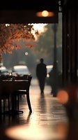 Atmospheric evening restaurant scene with silhouette of lone person walking past. Warm lighting, autumn leaves, and wine glass on table create moody ambiance.