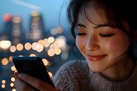 Young Asian woman smiling while using smartphone at night, city lights bokeh in background, wearing cozy sweater, natural makeup and red lipstick.