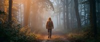 Young woman hiker walking alone on misty forest trail during golden hour autumn morning. Atmospheric fog creates dreamy backlit scene with warm sunlight filtering through trees.