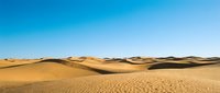 Vast golden sand dunes stretch across arid desert landscape under clear blue sky, showcasing natural terrain patterns and sandy wilderness environment.