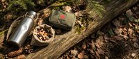 Outdoor camping gear with thermos bottle and metal container on wooden log surrounded by pine branches and autumn leaves in forest setting.