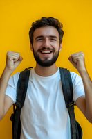 Young bearded man celebrating success with raised fists against vibrant yellow wall, wearing white t-shirt and backpack, expressing joy and achievement.