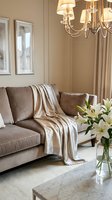 Elegant living room interior featuring a taupe upholstered sofa with decorative pillows and a draped throw blanket, crystal chandelier, framed artwork, flowing curtains, fresh white lilies in a glass vase, and marble coffee table for home decor inspiration.