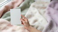 Female hand holding blank white card on soft pastel blankets and textiles, suitable for greeting cards, stationery mockups, and lifestyle marketing campaigns.