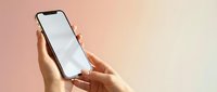 Hands holding smartphone with blank white screen against soft pink and beige gradient background for app mockup and mobile interface design.