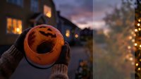 Hands in knitted gloves holding orange jack-o-lantern mug with hot beverage during Halloween evening with warm house lights and autumn decorations in background.