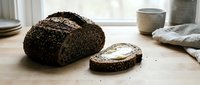 Freshly baked whole grain bread loaf with butter spread on slice, displayed on wooden table with kitchen items and natural window light.