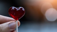Red heart shaped lollipop held between fingers against blurred background with warm bokeh lights, romantic sweet treat for Valentine's Day celebration.
