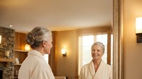 Two mature Caucasian women in white bathrobes enjoying spa relaxation time in luxury wellness center with warm lighting and stone fireplace.