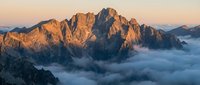 Dramatic mountain peaks emerging from sea of clouds at golden sunrise, alpine landscape with warm light illuminating rocky summits and misty valleys below.