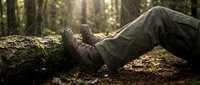 Hiker resting legs on fallen log in sunlit forest during outdoor adventure trek. Worn leather boots and cargo pants suggest experienced wilderness explorer.
