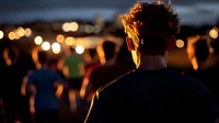 Young man with curly hair watching outdoor festival at dusk, silhouettes of crowd against blurred bokeh lights creating atmospheric evening ambiance.